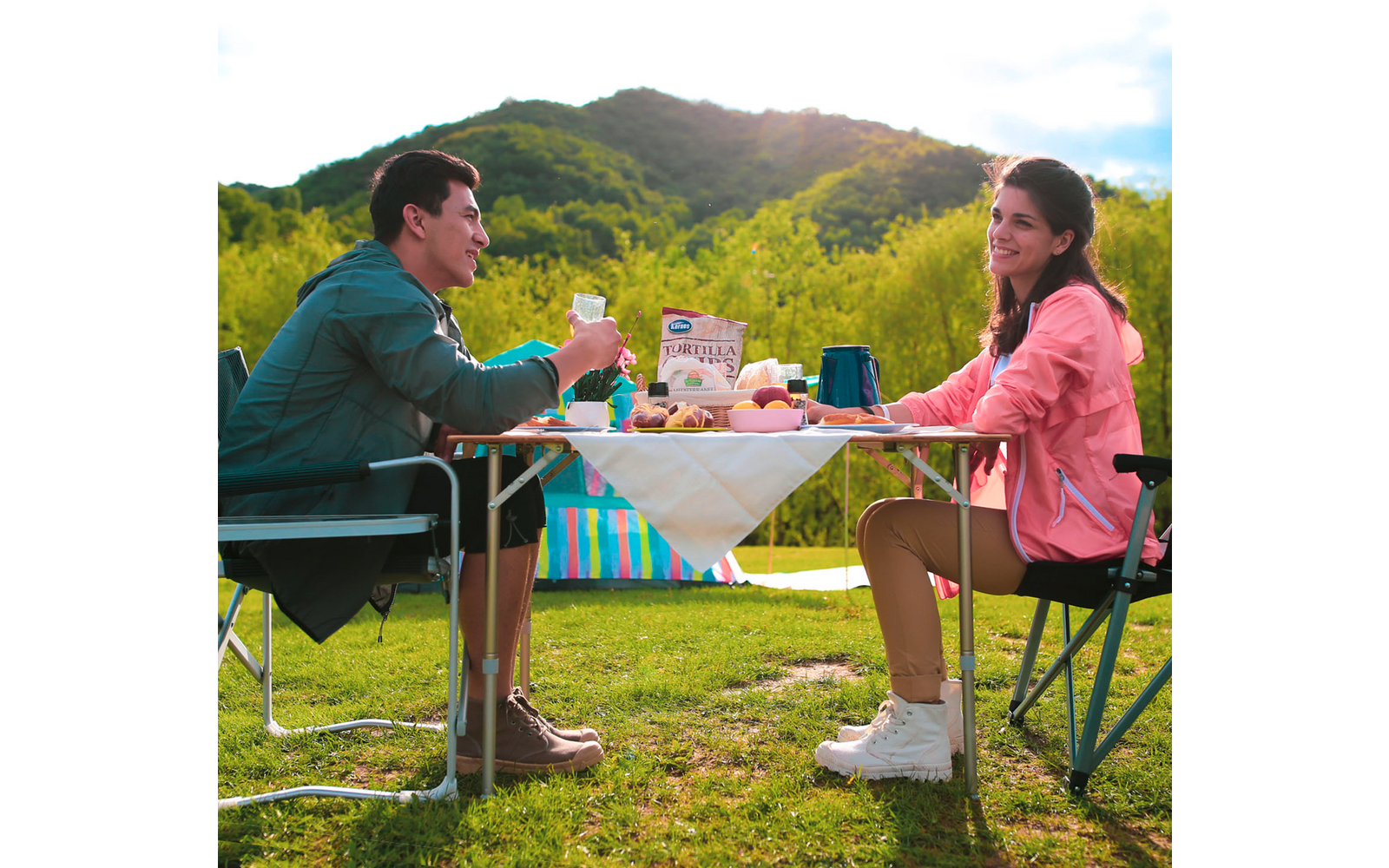 Deux personnes assises à une table en plein air avec des boissons et de la nourriture, une colline boisée au coucher du soleil en arrière-plan