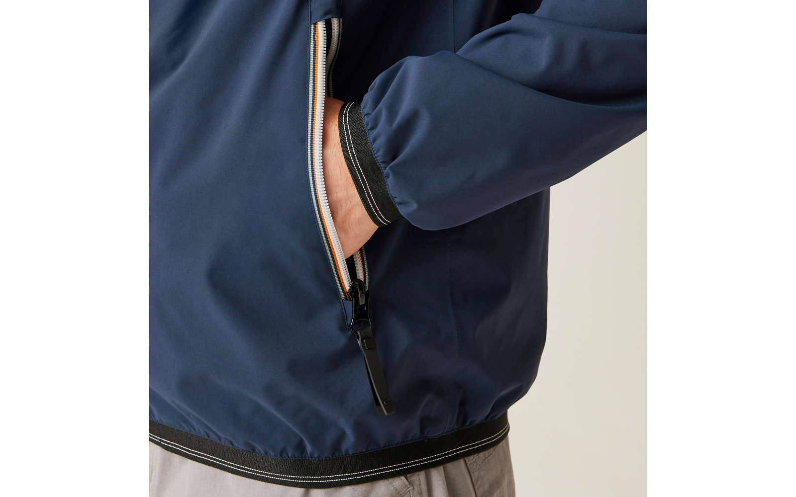 Nahaufnahme einer dunkelblauen Jacke mit zippbarer Seitentasche, in die eine Hand gesteckt ist; sichtbarer Reißverschluss mit mehrfarbigem Besatz und schwarzem Zipper, elastischer Saum und Ärmelbund, graue Hose sichtbar