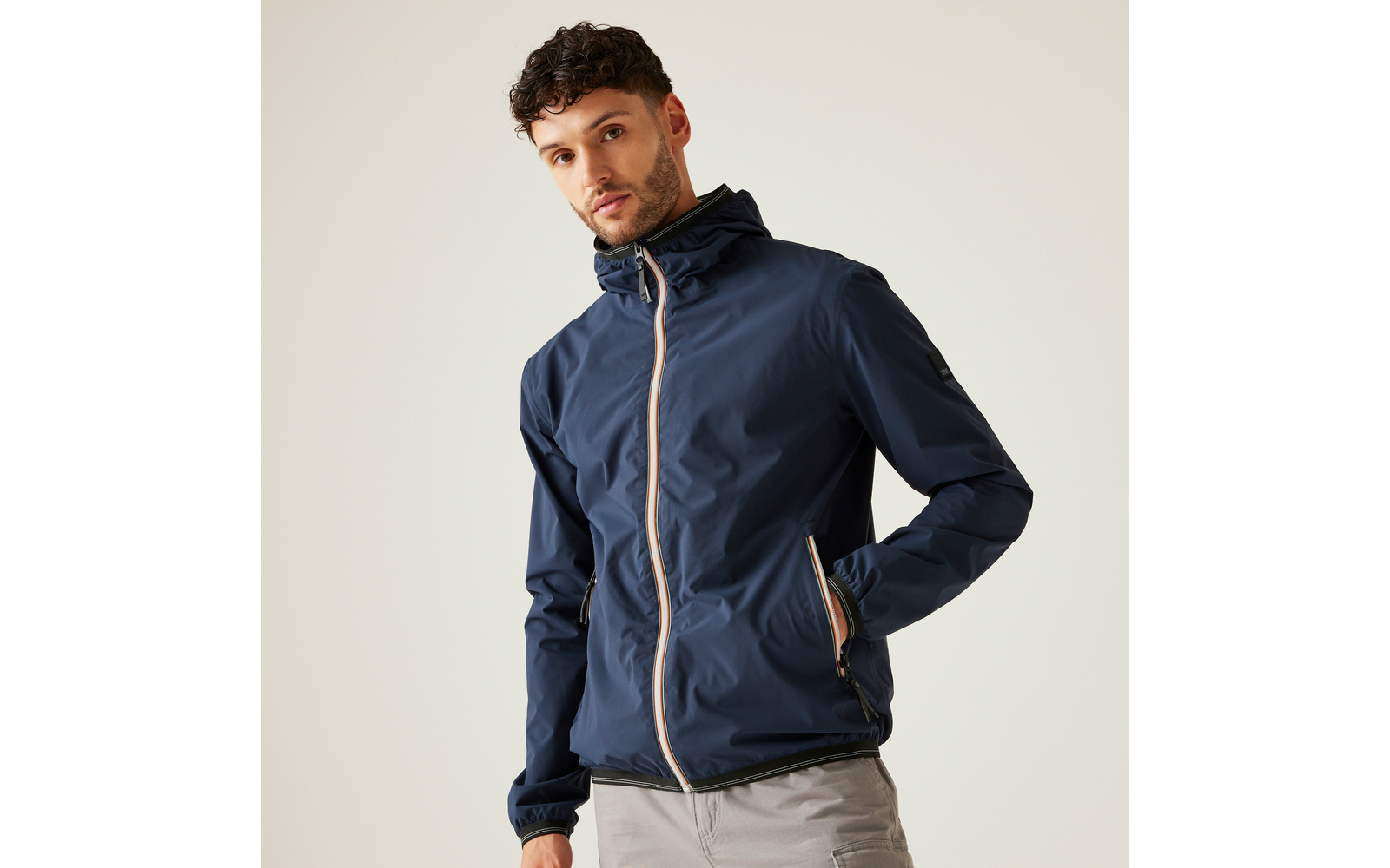 Mann mit dunklem, lockigem Haar und kurzem Bart trägt eine dunkelblaue Kapuzenjacke mit hellen Reißverschlusskanten und hellgraue Hosen vor hellem Hintergrund