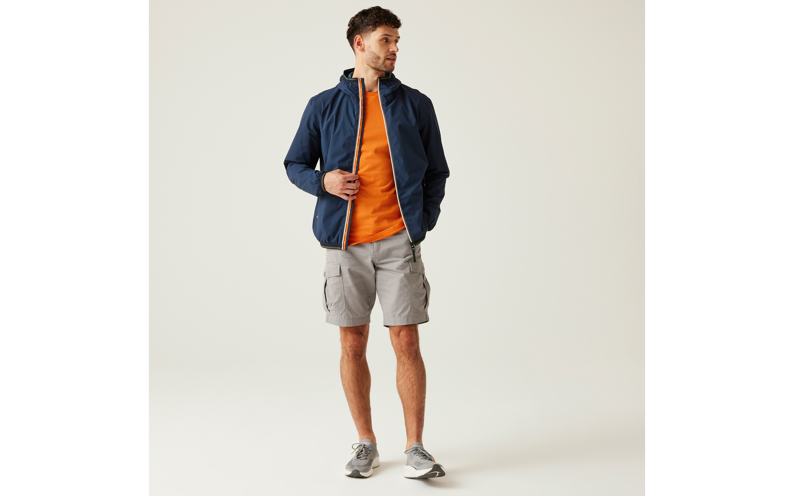 Person steht vor hellem Hintergrund, trägt dunkelblaue Jacke mit Kapuze, orangefarbenes T‑Shirt, hellgraue Cargo‑Shorts und graue Turnschuhe, Blick nach links