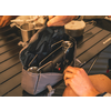 Graue offene Tasche auf einem dunklen, gerillten Tisch, darin zwei schwarze Gegenstände mit runden Metallgittern, zwei Hände halten die Tasche, im Hintergrund ein Metallbecher und ein Topf