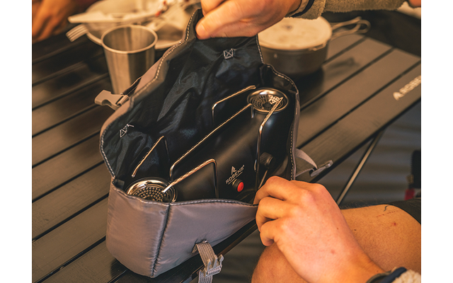 Graue offene Tasche auf einem dunklen, gerillten Tisch, darin zwei schwarze Gegenstände mit runden Metallgittern, zwei Hände halten die Tasche, im Hintergrund ein Metallbecher und ein Topf