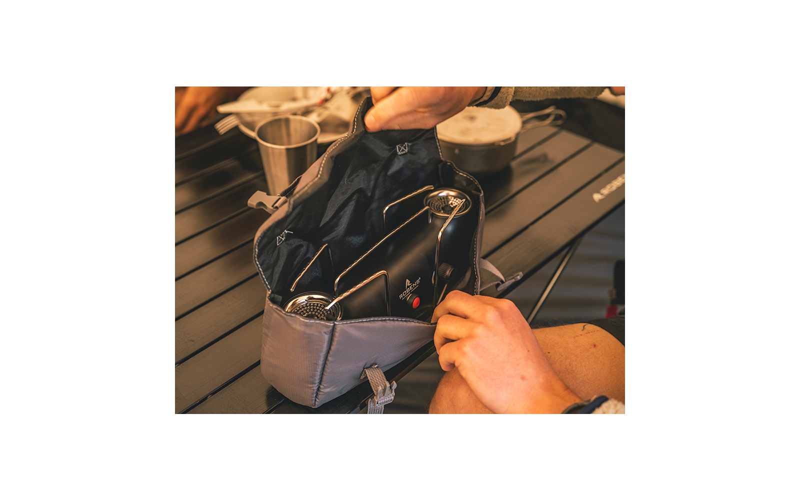Graue offene Tasche auf einem dunklen, gerillten Tisch, darin zwei schwarze Gegenstände mit runden Metallgittern, zwei Hände halten die Tasche, im Hintergrund ein Metallbecher und ein Topf