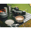 Schwarzer klappbarer Campingtisch mit tragbarem Gaskocher und Gaskartusche, grauer Teekessel mit orangenem Griff, Metallbecher mit Besteck, offener Topf mit hellem Inhalt, Deckel und Löffel, grünes Zelt im Hintergrund