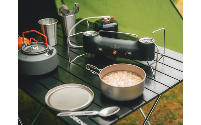 Schwarzer klappbarer Campingtisch mit tragbarem Gaskocher und Gaskartusche, grauer Teekessel mit orangenem Griff, Metallbecher mit Besteck, offener Topf mit hellem Inhalt, Deckel und Löffel, grünes Zelt im Hintergrund