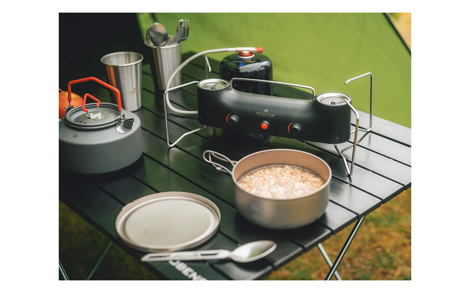 Schwarzer klappbarer Campingtisch mit tragbarem Gaskocher und Gaskartusche, grauer Teekessel mit orangenem Griff, Metallbecher mit Besteck, offener Topf mit hellem Inhalt, Deckel und Löffel, grünes Zelt im Hintergrund