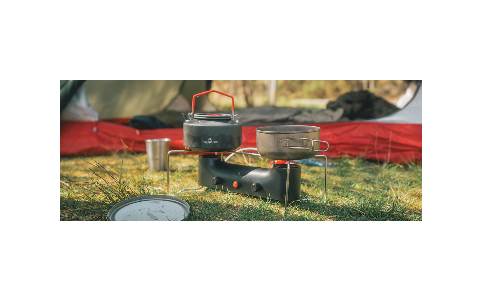 Tragbarer Gaskocher mit Teekessel, Topf, Metallbecher und Deckel auf Gras vor einem Zelt