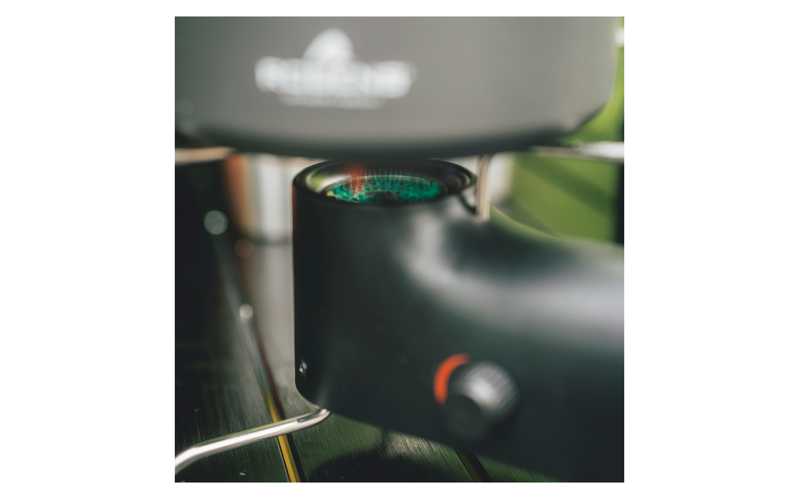 Nahaufnahme eines schwarzen Teils einer Kaffeemaschine mit rundem Metallgitter, grünen Lichtreflexen, schwarzem Gehäuse, silberner Metallstange und schwarzem Drehknopf mit orangem Ring