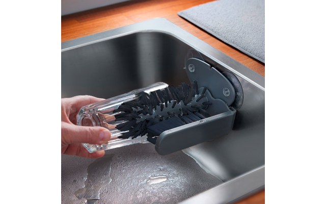 Mano sosteniendo un vaso transparente colocado en un cepillo gris con ventosa sujeto al fregadero de acero inoxidable; agua en el fregadero y una alfombrilla gris sobre la encimera de madera
