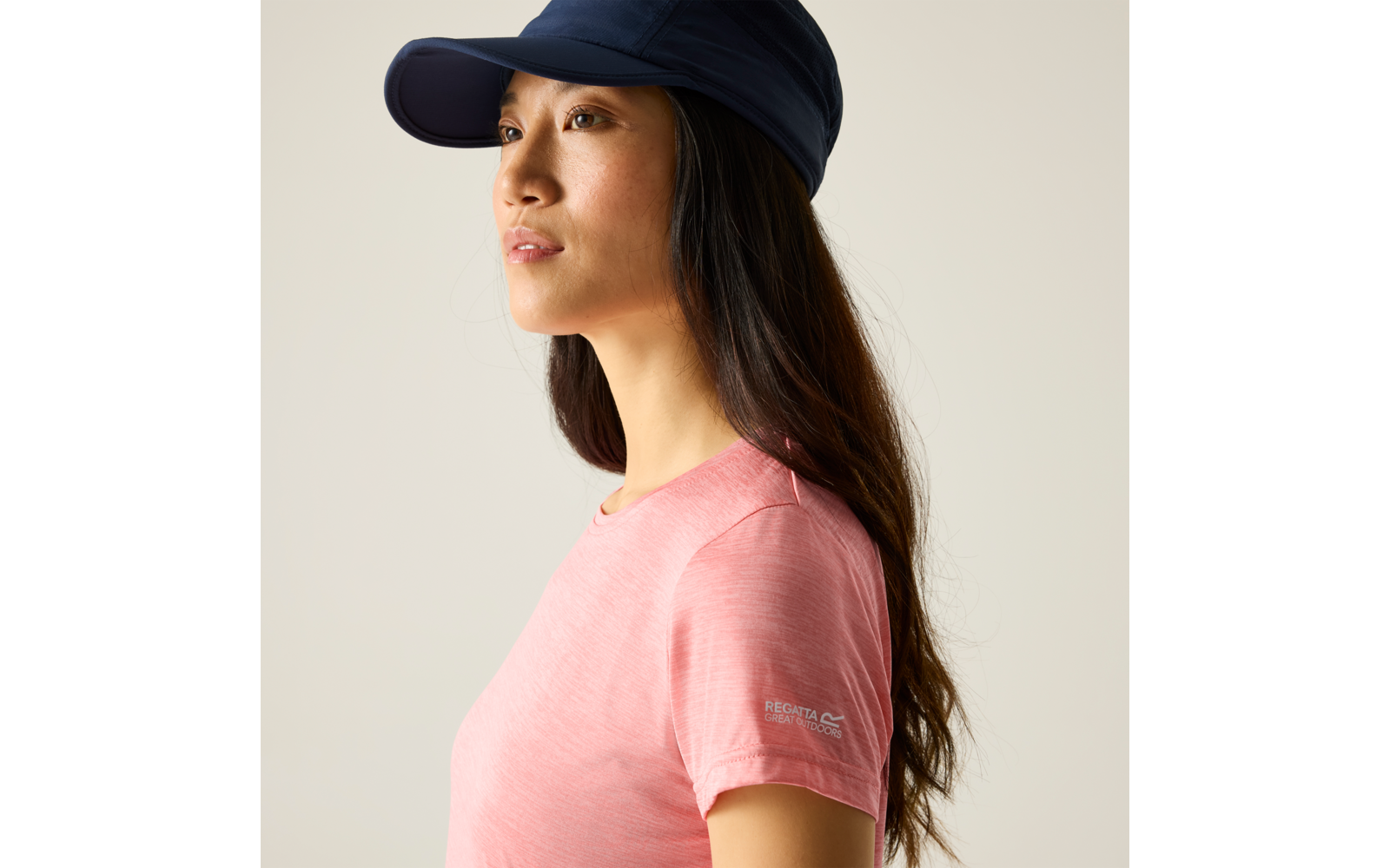 Profiel van een persoon met lang donker haar, een donkerblauwe pet en een roze T‑shirt met een klein logo op de mouw, effen lichte achtergrond