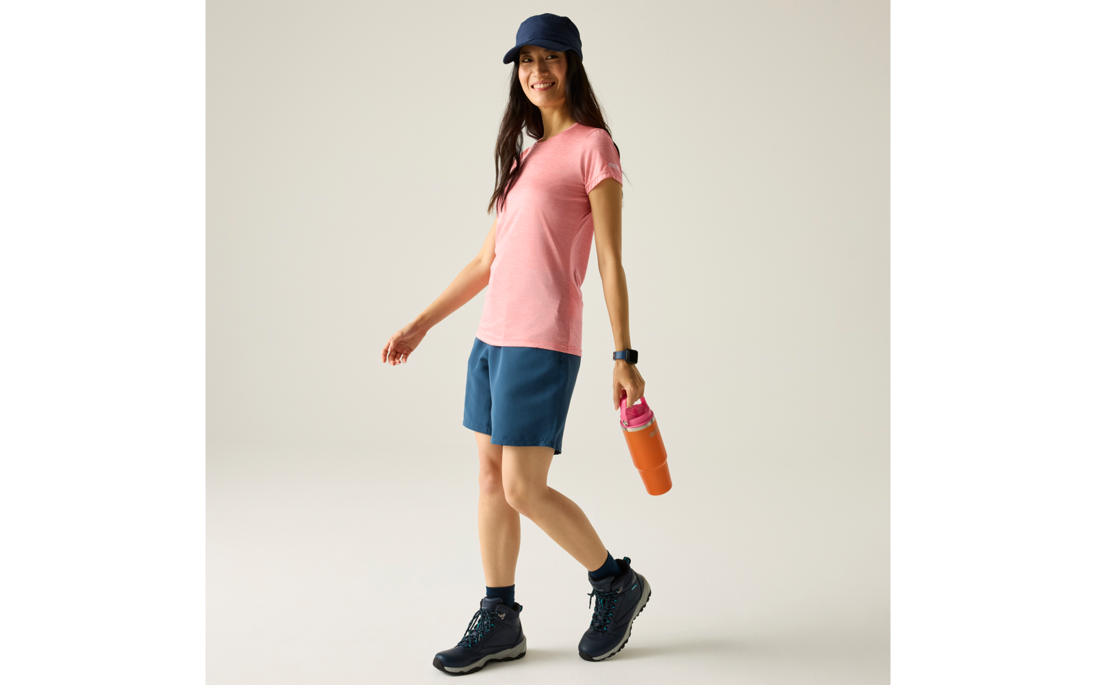 Persoon met roze T-shirt, blauwe korte broek en donkerblauwe pet, houdt een oranje drinkfles met roze dop vast, draagt wandelschoenen en een smartwatch, lopend voor een lichte achtergrond