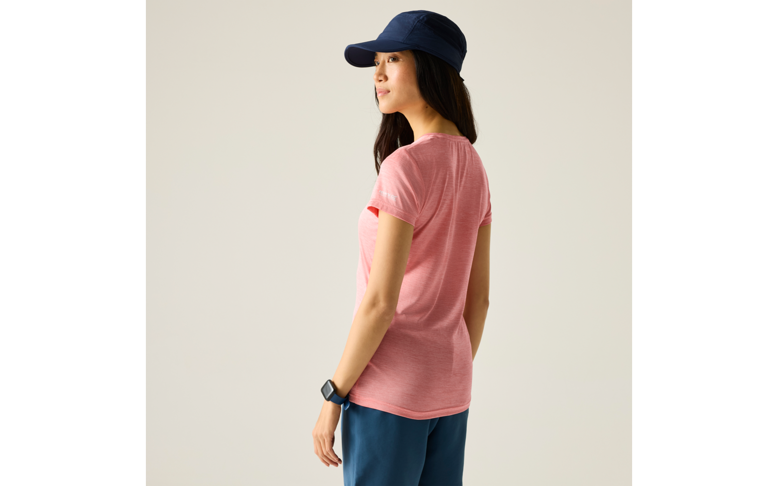 Persoon in roze T‑shirt, donker lang haar, donkerblauwe pet, donkerblauwe broek, smartwatch om linkerpols, iets weggedraaid tegen een lichte achtergrond