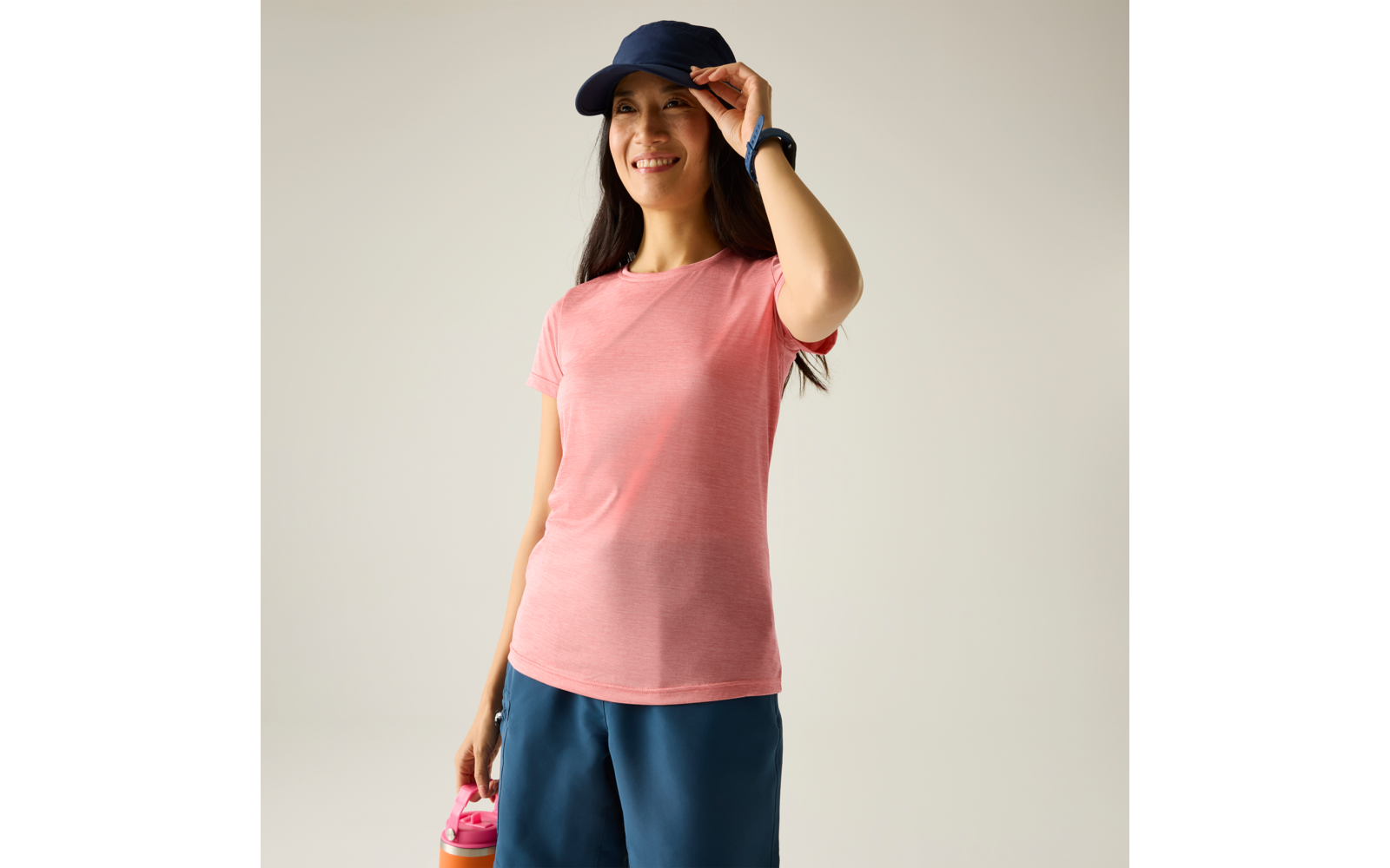 Vrouw in roze T-shirt en donkerblauwe pet, houdt een roze drinkfles vast, draagt blauwe shorts en een polshorloge, effen lichte achtergrond