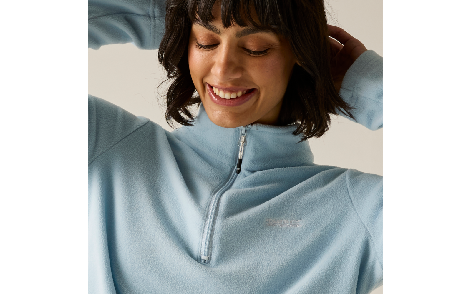 Persona sorride con occhi chiusi, indossa un pile azzurro chiaro con cerniera corta, mani dietro la testa, capelli scuri con frangia, sfondo uniforme