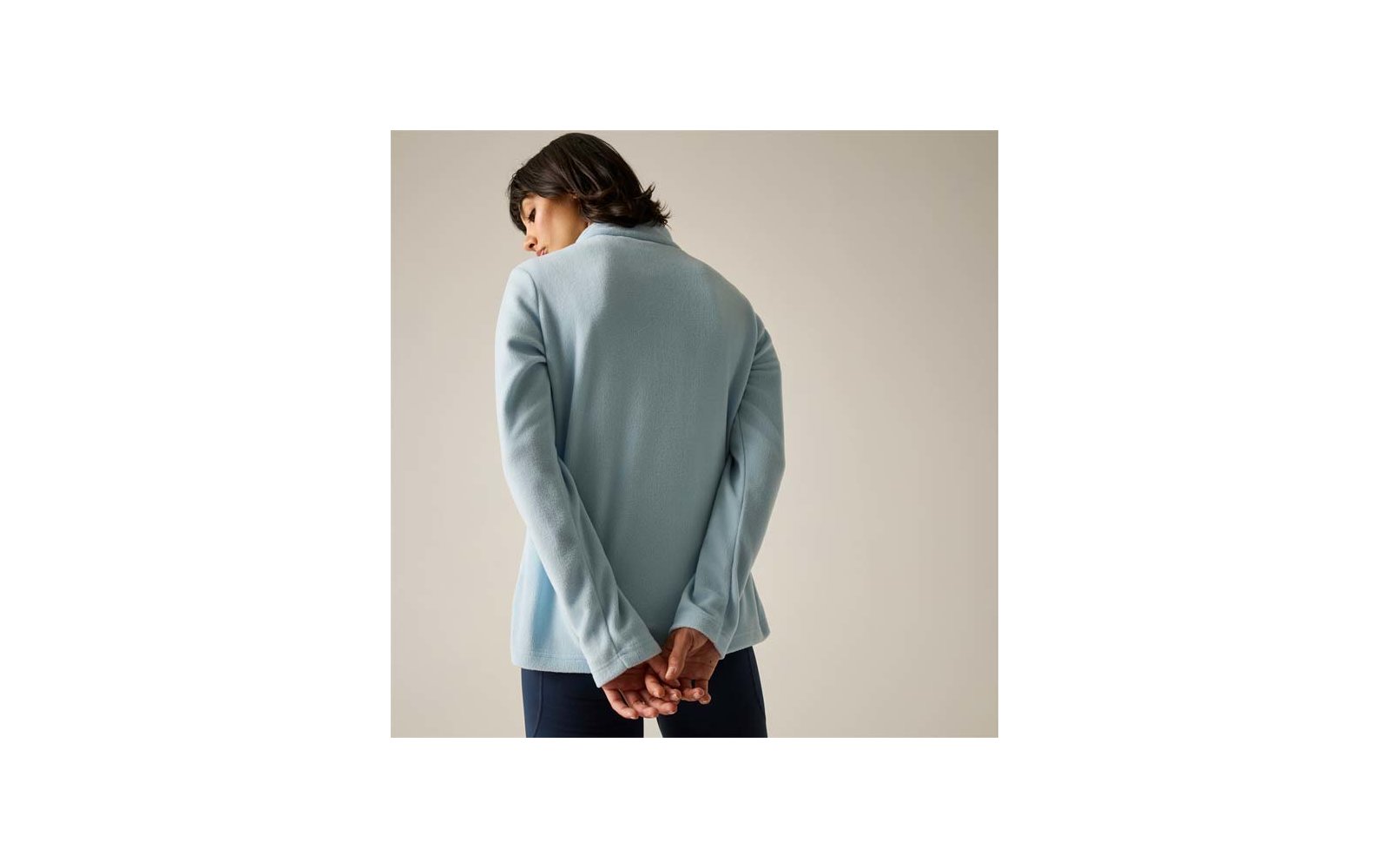 Vista posteriore di una persona con capelli corti e scuri che indossa un capo manica lunga azzurro chiaro con collo alto e pantaloni scuri, mani intrecciate dietro la schiena, su sfondo neutro chiaro