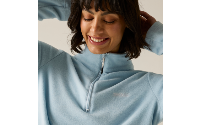 Persona sorride con occhi chiusi, indossa un pile azzurro chiaro con cerniera corta, mani dietro la testa, capelli scuri con frangia, sfondo uniforme