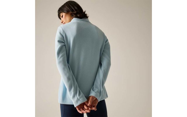 Vista posteriore di una persona con capelli corti e scuri che indossa un capo manica lunga azzurro chiaro con collo alto e pantaloni scuri, mani intrecciate dietro la schiena, su sfondo neutro chiaro
