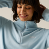 Persona sorride con occhi chiusi, indossa un pile azzurro chiaro con cerniera corta, mani dietro la testa, capelli scuri con frangia, sfondo uniforme