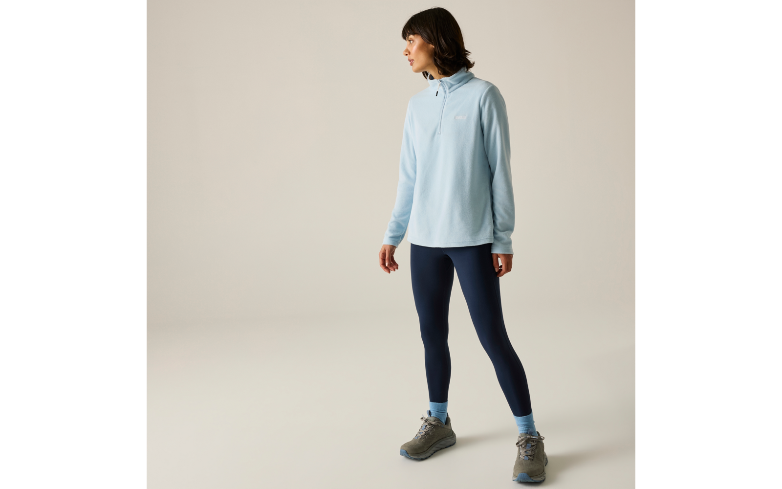 Persona in profilo su sfondo chiaro, indossa un maglione azzurro con mezza cerniera, leggings blu scuro, calzini azzurri e scarpe da trekking grigie