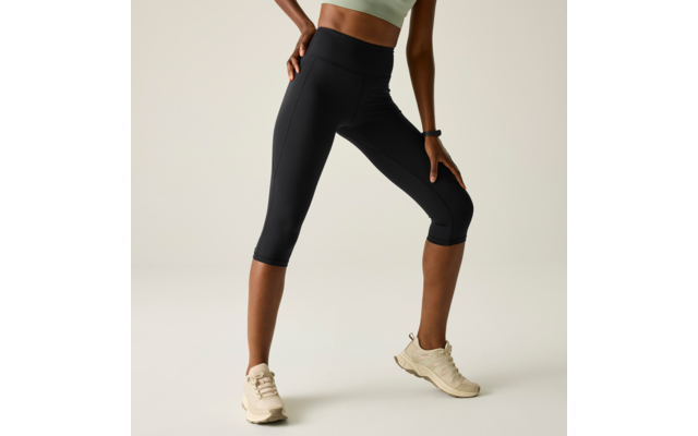 Lower body of a standing person wearing black knee-length leggings, a light green crop top, and beige sneakers; hands on hip and thigh; plain light background