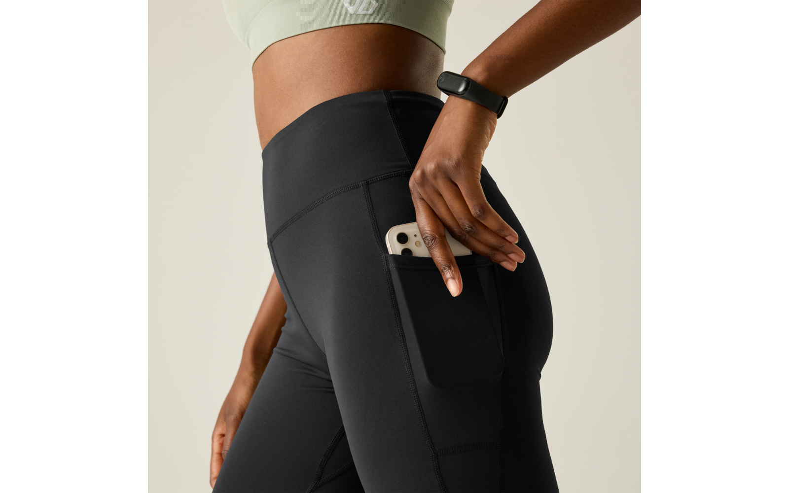 Close-up of a person from waist to thighs wearing black athletic leggings with a side pocket, placing a smartphone into the pocket; light green sports top and fitness tracker on the wrist visible