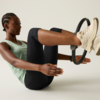 Person seated in a V-sit, holding a Pilates ring between their feet, wearing a tank top, leggings and white sneakers against a light background