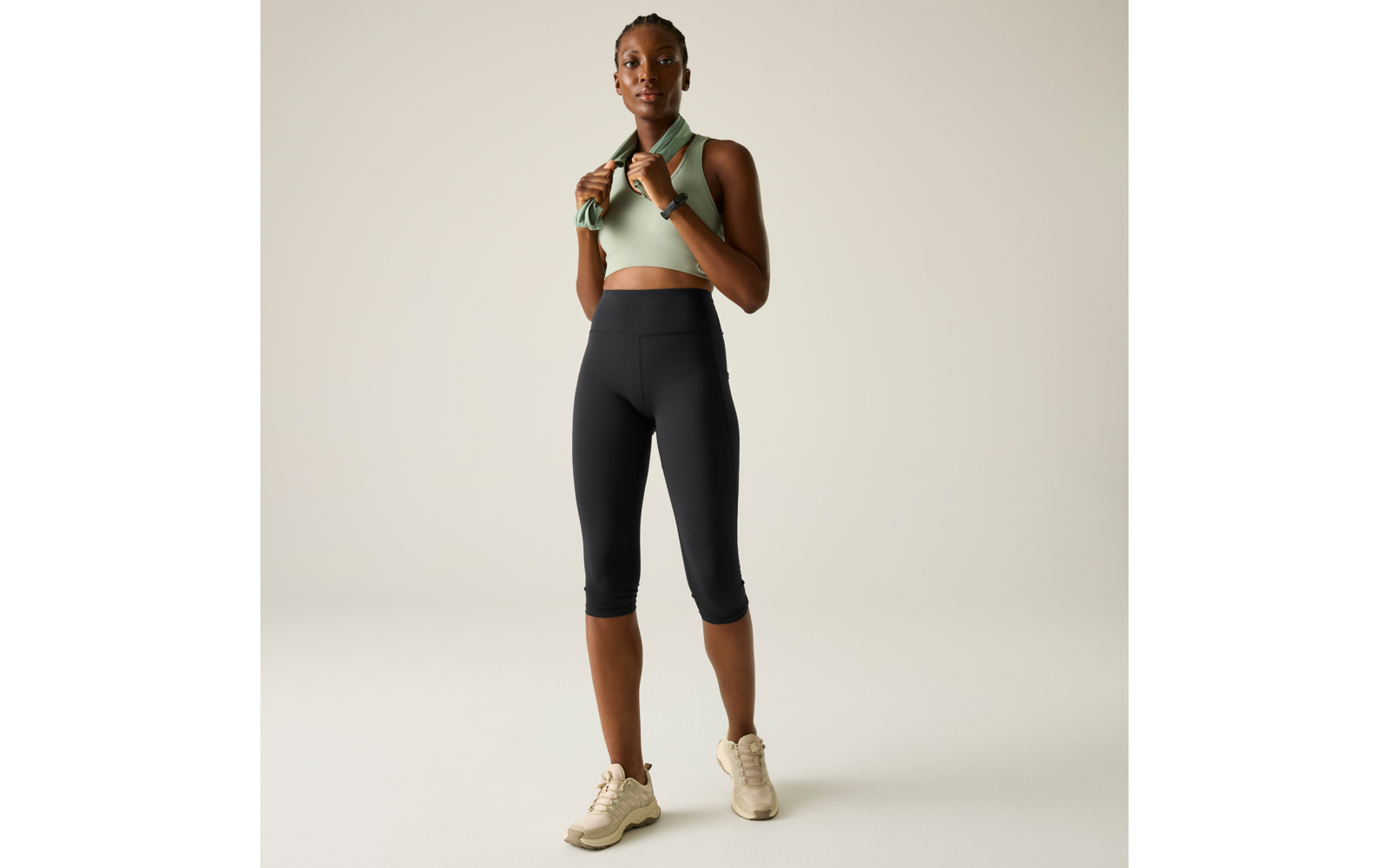 Person standing against a light background wearing a mint sports crop top, black capri leggings, beige sneakers and a towel around the neck