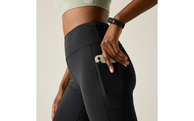 Close-up of a person from waist to thighs wearing black athletic leggings with a side pocket, placing a smartphone into the pocket; light green sports top and fitness tracker on the wrist visible