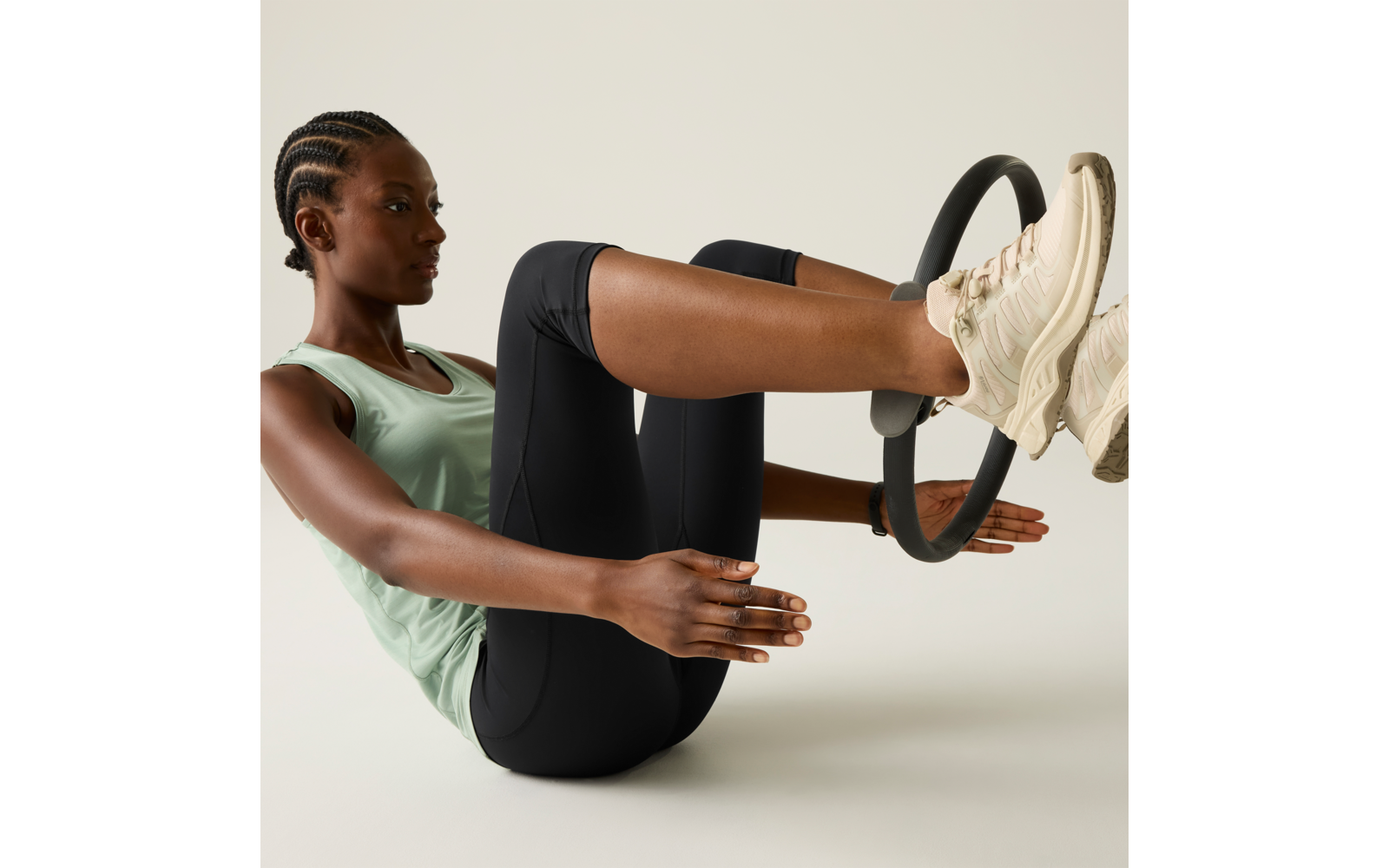 Person seated in a V-sit, holding a Pilates ring between their feet, wearing a tank top, leggings and white sneakers against a light background