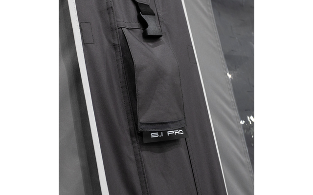 Nahaufnahme eines dunklen Stoffpaneels mit vertikalem Reißverschluss, Riemen, kleinem Label 'S.I PRO', weißen Paspeln und glänzender transparenter Plane rechts