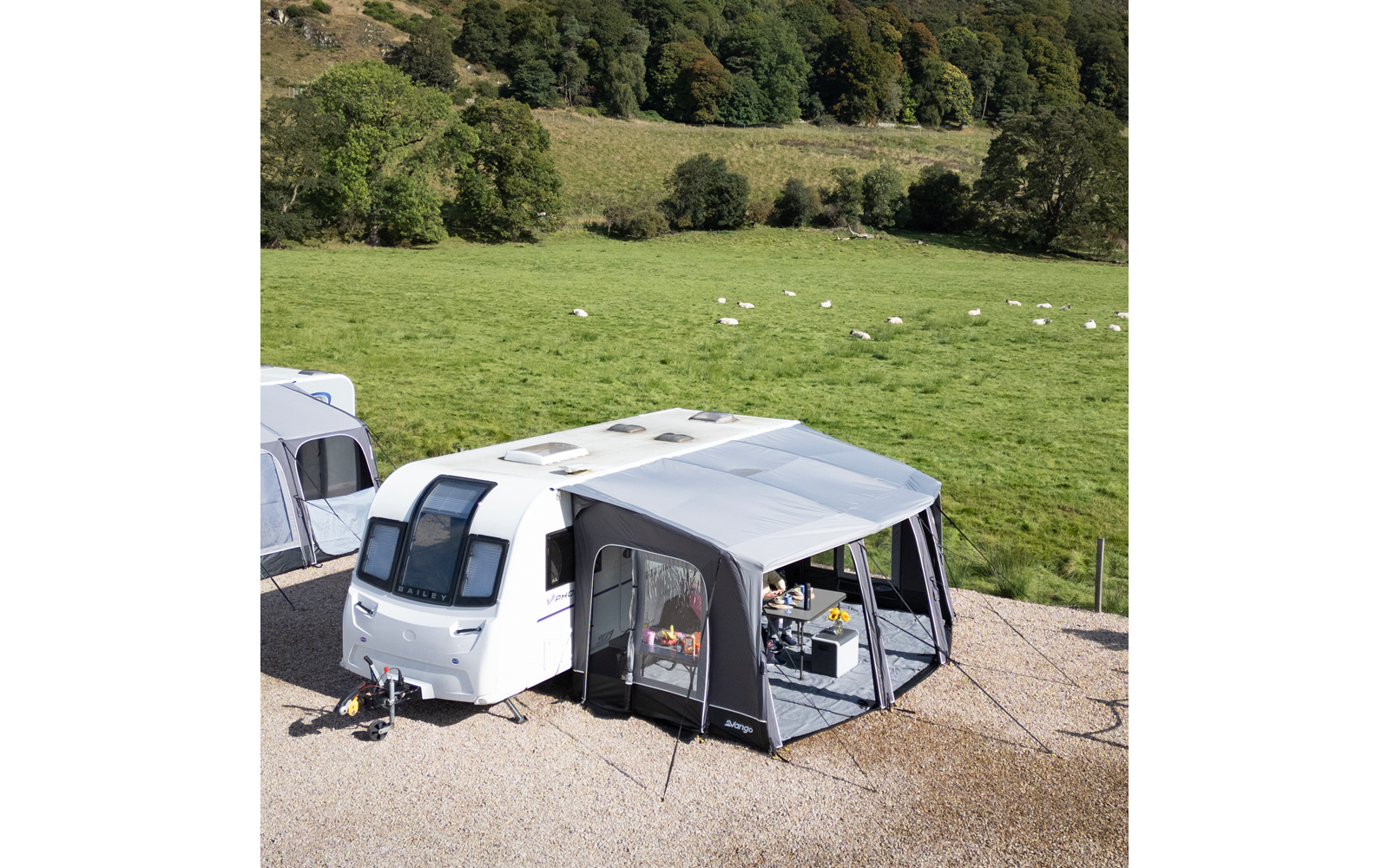 Weißer Wohnwagen mit grauem Vorzelt auf Schotter; im Vorzelt ein Tisch mit Gegenständen; im Hintergrund grüne Wiese mit Schafen und Bäumen