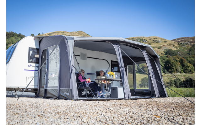 Wohnwagen mit angebautem Vorzelt; zwei sitzende Personen an einem Tisch im Vorzelt; Tassen und Vase mit Blumen auf dem Tisch; Schotterboden; grüne Hügel und blauer Himmel im Hintergrund