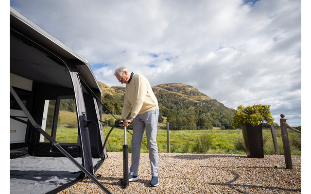 Mann steht auf Kies neben einem Wohnwagenvorzelt und hält eine schwarze Handpumpe; im Hintergrund Berg, Wiese und bewölkter Himmel