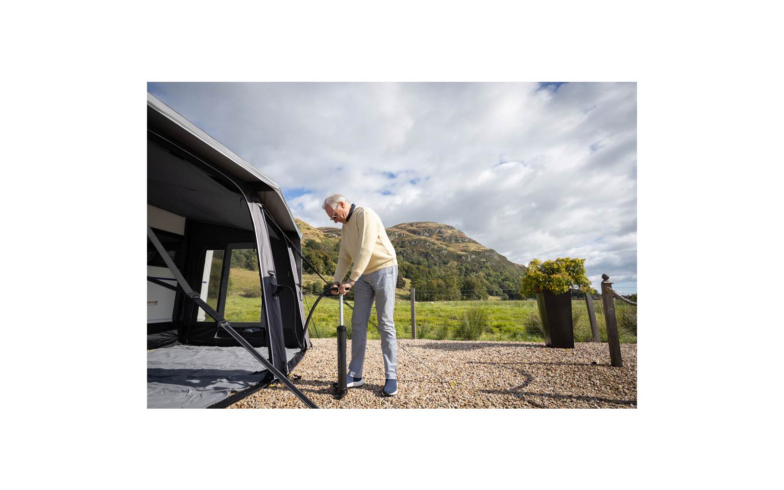 Mann steht auf Kies neben einem Wohnwagenvorzelt und hält eine schwarze Handpumpe; im Hintergrund Berg, Wiese und bewölkter Himmel