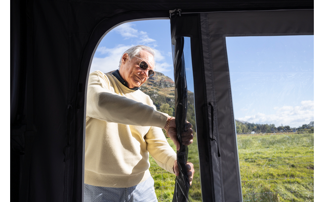 Mann mit grauem Haar und Sonnenbrille zieht den Reißverschluss einer Stofftür auf; Wiese und Hügel im Hintergrund