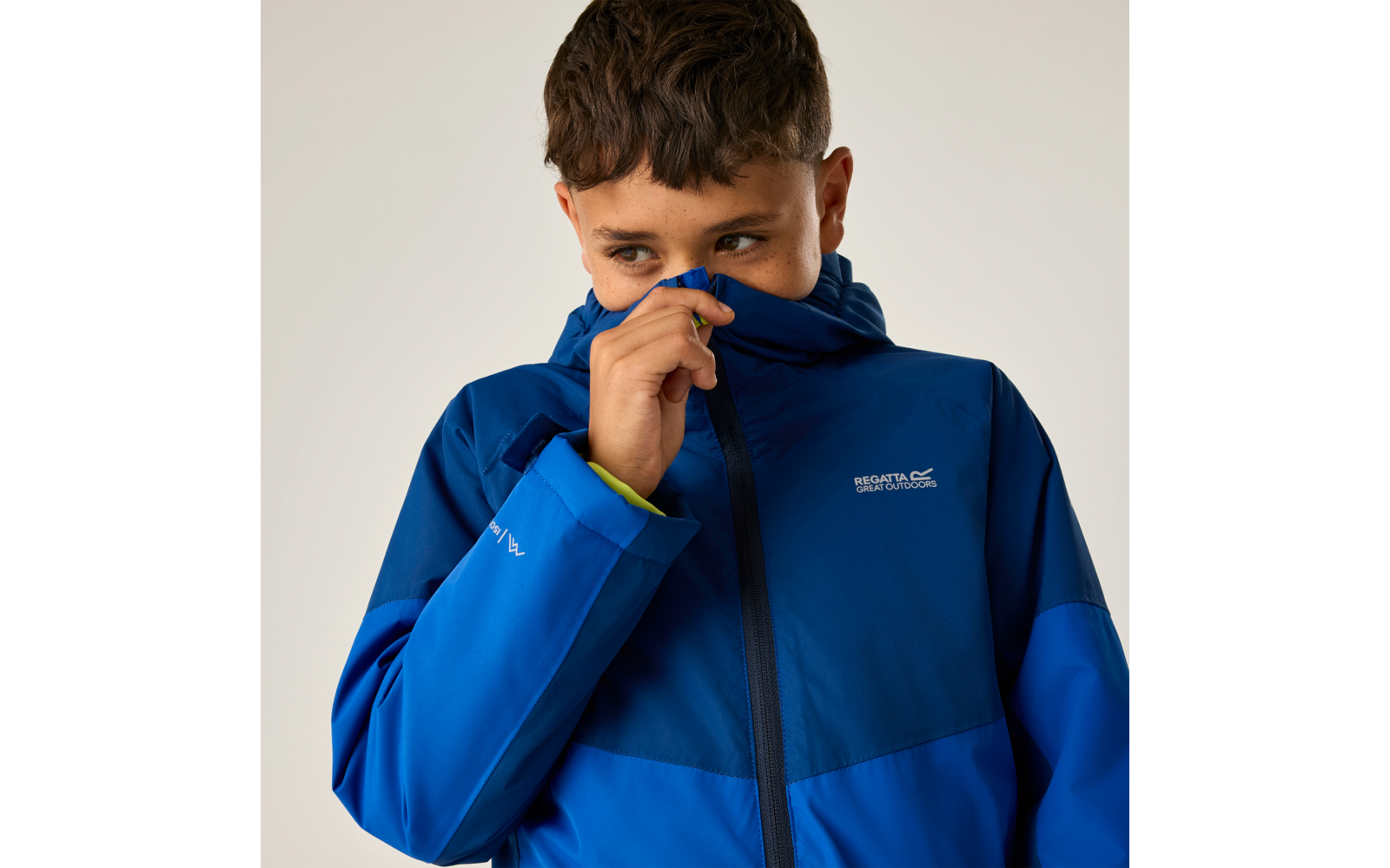 Junge in blauer Kapuzenjacke zieht Reißverschluss bis zur Nase, Blick nach rechts, heller Hintergrund