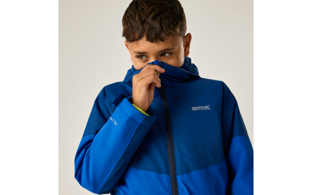Junge in blauer Kapuzenjacke zieht Reißverschluss bis zur Nase, Blick nach rechts, heller Hintergrund