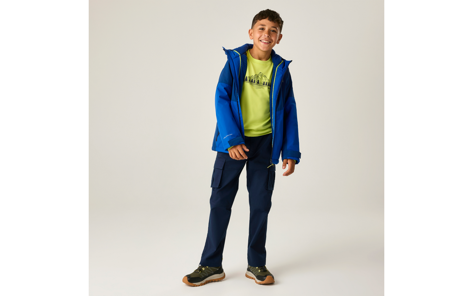 Eine lächelnde Person steht vor hellem Hintergrund, trägt eine blaue Jacke, ein neongrünes T‑Shirt, dunkle Cargohosen und Sportschuhe