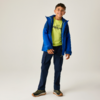 Eine lächelnde Person steht vor hellem Hintergrund, trägt eine blaue Jacke, ein neongrünes T‑Shirt, dunkle Cargohosen und Sportschuhe