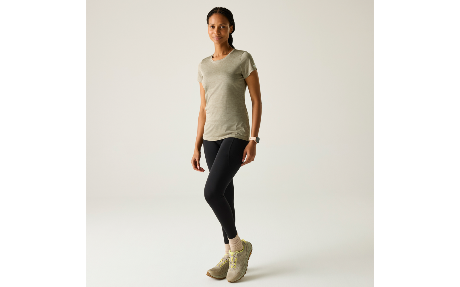 Persoon staat voor een lichte achtergrond, draagt een olijfgroen T‑shirt, zwarte legging, lichte sokken en groenachtige sportschoenen, smartwatch om linkerpols, haar naar achteren gebonden