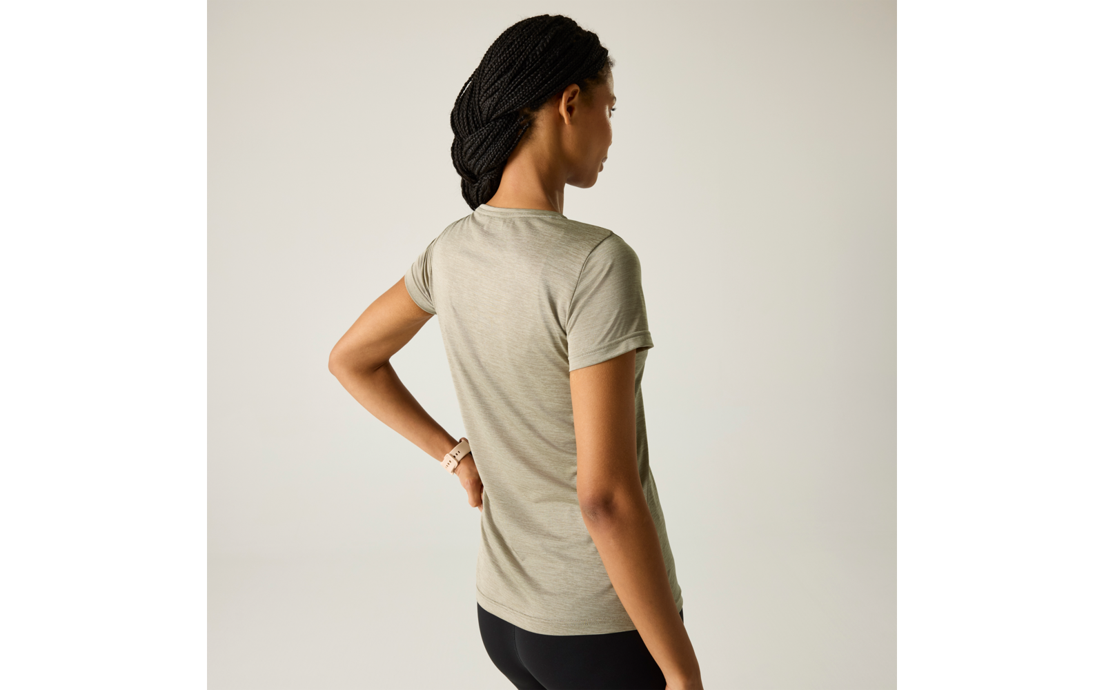 Persoon van achteren met lichtolijfgroen T‑shirt en zwarte legging, ingevlochten haar, één hand op de heup, lichte achtergrond