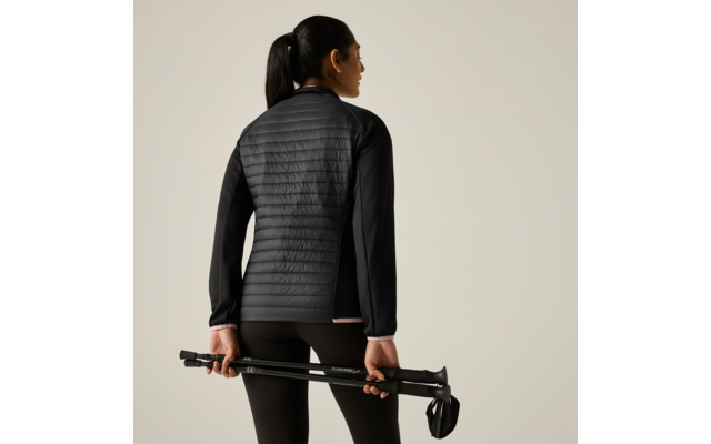 Persona vista da dietro con giacca nera trapuntata e leggings, tiene due bastoncini da trekking ripiegati dietro la schiena, sfondo chiaro uniforme