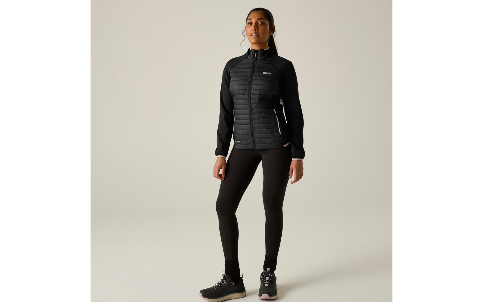 Persona in piedi, rivolta in avanti, indossa giacca nera trapuntata con cerniera, leggings neri e scarpe sportive nere, sfondo chiaro uniforme