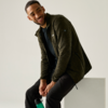 Ein Mann sitzt auf einem weißen Block, trägt eine dunkelgrüne Reißverschlussjacke, schwarzes Shirt und schwarze Hosen, hält eine grüne Trinkflasche, lächelt und schaut seitlich