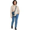 Persoon staand voor een witte achtergrond met een beige fleecejack, wit shirt, blauwe spijkerbroek, zwarte sneakers en een kleine blauwe crossbodytas, kijkt in de camera en lacht