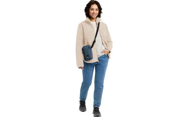 Persoon staand voor een witte achtergrond met een beige fleecejack, wit shirt, blauwe spijkerbroek, zwarte sneakers en een kleine blauwe crossbodytas, kijkt in de camera en lacht