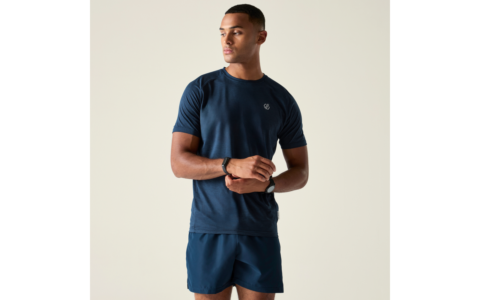 Homme en T-shirt et short bleu foncé, debout légèrement de profil, mains jointes devant la taille, regard tourné vers la gauche
