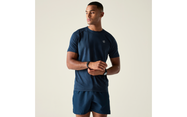 Homme en T-shirt et short bleu foncé, debout légèrement de profil, mains jointes devant la taille, regard tourné vers la gauche