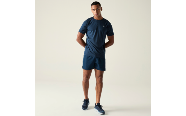 Homme debout en t‑shirt bleu foncé, short bleu foncé et baskets sur fond clair, mains derrière le dos