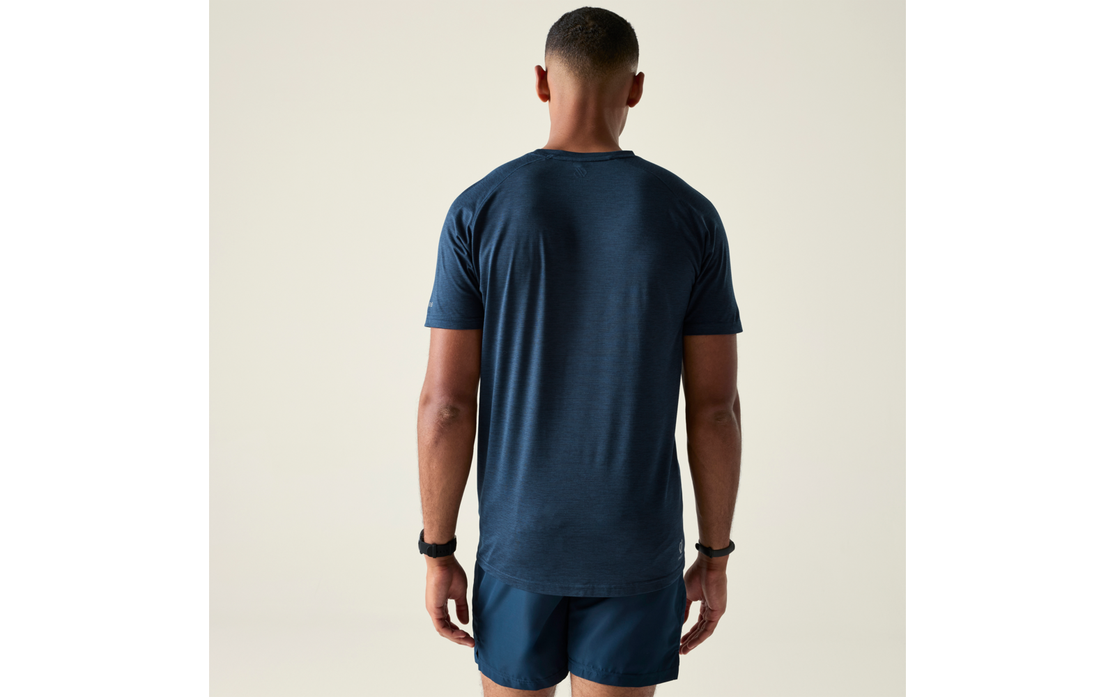 Homme vu de dos, portant un t-shirt bleu foncé et un short bleu foncé, debout devant un fond clair uni